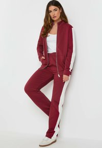 Tuta bordeaux con giacca zip e strisce bianche sui lati, abbinata a pantaloni. Il tessuto sembra morbido con una vestibilità casual.
