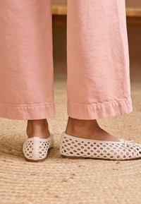 Bailarinas tejidas blancas con punta redonda y detalles de encaje, combinadas con pantalones de tiro ancho en un suave tono rosa. Superficie texturizada sobre una alfombra de fibra natural.