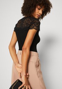 Body negro acanalado con un yugo de encaje, que presenta bordes con forma de concha. Combinado con pantalones beige de corte a medida, los accesorios incluyen un clutch negro.