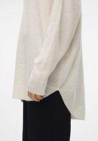 Cream-colored sweater with a ribbed hem and side slits, featuring a soft texture and long sleeves. Paired with black pants.