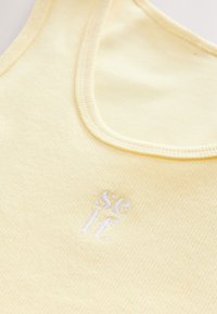 Débardeur jaune côtelé avec encolure ronde et texte brodé blanc "se" positionné en bas à gauche. Texture douce.