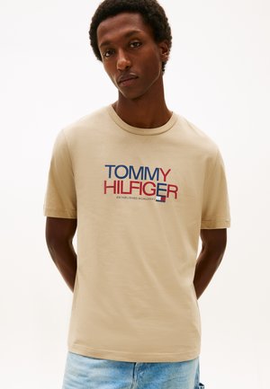 Jonge man draagt een beige Tommy Hilfiger T-shirt met rood en blauw logo, staand tegen een effen witte achtergrond.