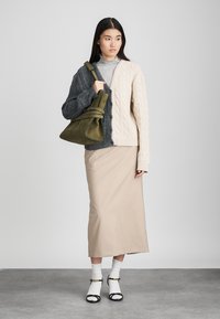 Πλεγμένο καρδιοκάλυμμα σε γκρίζο και κρεμ, ραμμένη μακριά φούστα σε μπεζ, πράσινη τσάντα, άσπρες κάλτσες και μαύρες σανδάλες με λουριά, σε ουδέτερο φόντο.