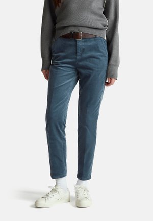 Personne portant un pantalon en velours côtelé bleu, des baskets blanches, des chaussettes blanches, un pull gris et une ceinture marron, debout sur fond blanc.