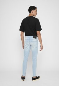 Jeune homme debout, dos tourné, portant un t-shirt noir, un jean skinny bleu clair et des baskets noires à semelles blanches.