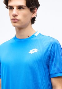 Νέος άνδρας που φοράει μπλε αθλητική μπλούζα με γεωμετρικά σχέδια και λευκό λογότυπο στο στήθος, στραμμένος ελαφρώς προς τα αριστερά μπροστά σε απλό φόντο.