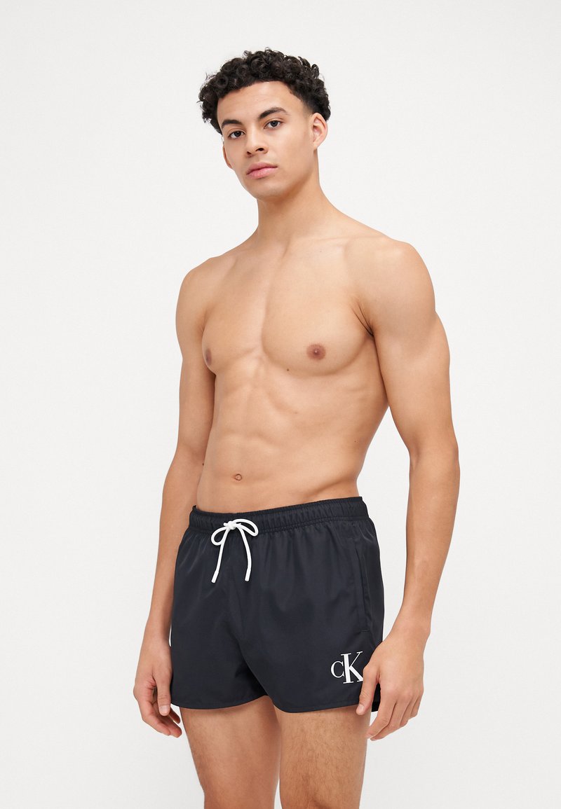 Shorts de bain noirs en tissu léger, dotés d'une taille élastique avec un cordon de serrage blanc et d'un petit logo "cK" blanc sur le côté.