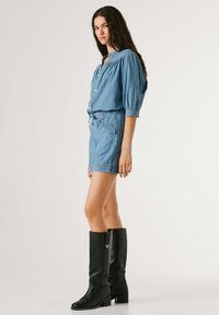 Mujer joven con un mono de manga larga a rayas azules y botas negras hasta la rodilla, de pie de lado contra un fondo liso.