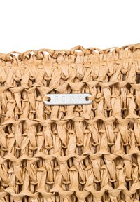 Geflochtener strohfarbenen Beutel in natürlichem Beige mit einem komplexen Muster. Ein metallisch silberner Anhänger zeigt "ROXY", in das Geflechtdesign eingestickt.