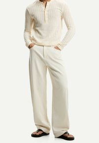 Suéter de mangas largas de color crema con un diseño acanalado y cuello tipo henley con botones, acompañado de pantalones anchos blancos y sandalias negras de dedo.