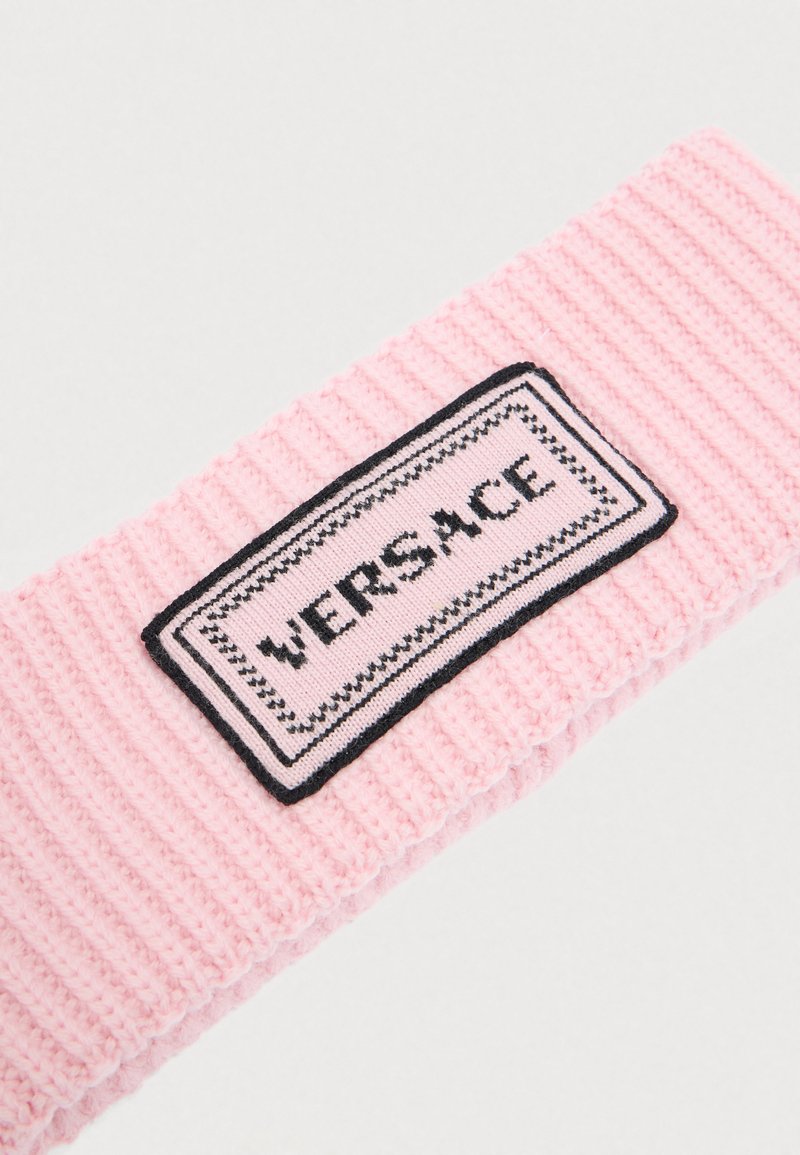 Pink knit headband with ribbed texture and a rectangular black patch featuring the "VERSACE" logo in bold letters and decorative borders.