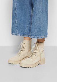 Beige ruskinds ankelstøvler med rund tå, snørresystem foran og en tekstureret gummisål, parret med brede denimbukser.