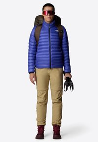 Giacca isolante blu, pantaloni khaki e stivali bordeaux. Il modello indossa uno zaino e tiene degli occhiali da sci. Le caratteristiche includono una texture gonfia e cerniere.