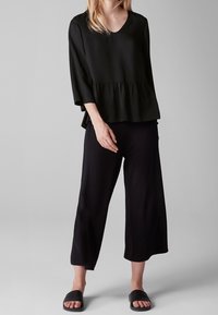 Schwarzes Peplum-Top mit Dreiviertelärmeln, kombiniert mit schwarzen weit geschnittenen Hosen und flachen schwarzen Sandalen, mit einem V-Ausschnitt.