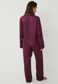 Satin pyjamasæt i dyb burgundy med en knaplukket top med krave og lange ærmer samt matchende bukser med elastisk talje.