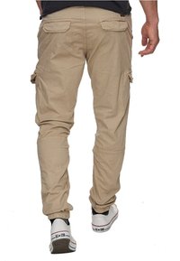 Homme portant un pantalon cargo beige et des baskets blanches marchant à l'écart, visible de la taille vers le bas sur un fond blanc.