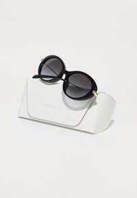 Ochelari de soare rotunzi negri cu lentile degrade, așezați pe o carcasă albă cu textură, care are inscripția „Michael Kors” embosată.