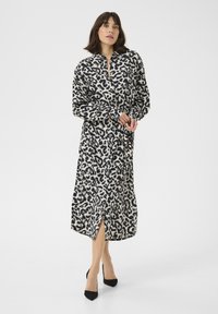 Žena u crno-bijeloj midi haljini s uzorkom leopardove kože, dugih rukava i s prorezom sprijeda, u crnim šiljastim cipelama na petu, stoji na bijeloj pozadini.