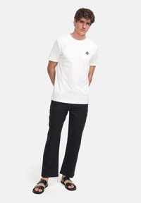 Vit bomull t-shirt med kort ärm och en liten svart logotyp, tillsammans med svarta byxor och svarta sandaler. Slät textur, enkel design.