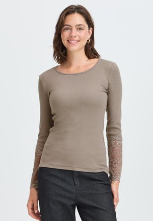 Jeune femme aux cheveux bruns mi-longs portant un haut taupe à manches longues avec des poignets en dentelle et un pantalon foncé, debout devant un fond clair.