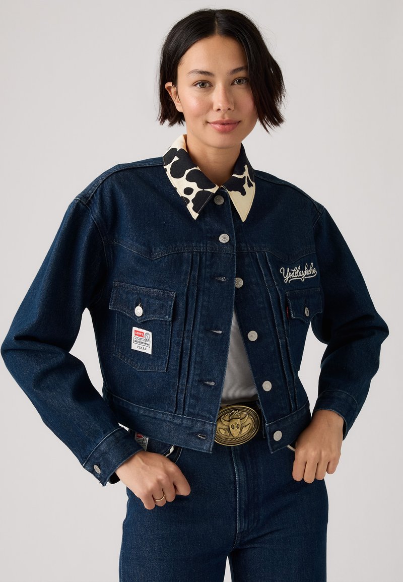 Giacca in denim blu scuro con colletto a stampa mucca in bianco e nero a contrasto, tasche frontali e dettagli ricamati sul petto.