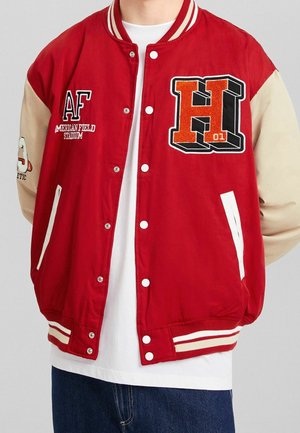 Rote und beige Collegejacke mit weißen Knöpfen, "H 01"-Aufnäher auf der Brust, Schriftzug "AF American Field Stadium", getragen über weißem Hemd und blauer Jeans.