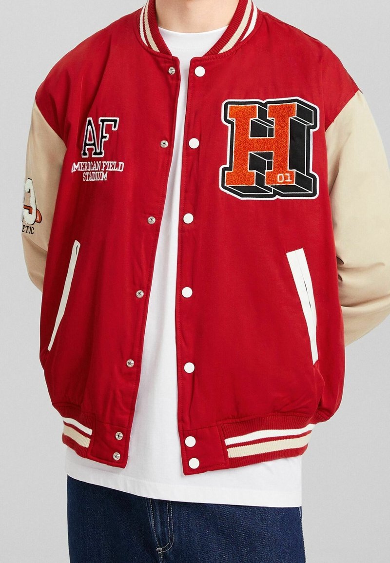 Veste universitaire rouge et beige avec boutons blancs, écusson "H 01" sur la poitrine, texte "AF American Field Stadium", portée sur une chemise blanche et un jean bleu.