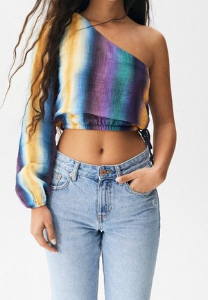 Femme portant un crop top asymétrique à manches longues avec des rayures verticales arc-en-ciel et un jean taille haute bleu clair, main dans la poche.
