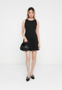 Femme jeune portant une robe noire sans manches, tenant un sac à main noir et portant des ballerines noires pointues, se tenant devant un fond blanc.