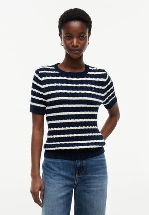 Person iført en navyblå og hvid stribet kortærmet ribstrikket sweater og blå jeans, stående med den ene hånd bag ryggen.