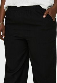 Juodos plačios kelnės, pagamintos iš tekstūruotos medžiagos, su elastinga juosmeniu ir dviem priekiniais kišenėmis funkcionalumui.