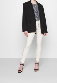 Černý oversized blazer, bílý top s malými námořnickými tečkami, krémové skinny džíny a černé sandály na vysokém podpatku. Jednoduchý detail s knoflíkem na džínech.