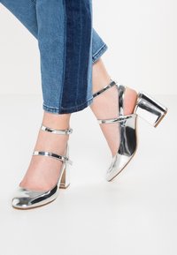 Piedi che indossano delle lucide mary jane argentate con tacco a blocco, abbinate a un paio di jeans blu a due tonalità, in piedi su un pavimento bianco.