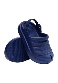 Ζευγάρι μπλε clogs χωρίς κορδόνια με υφασμένους ιμάντες και έναν ιμάντα γύρω από τη φτέρνα, εμφανίζονται με ένα παπούτσι όρθιο και το άλλο που δείχνει την σόλα.