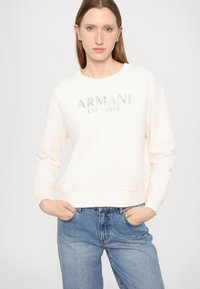 Felpa beige chiaro con polsini e orlo a costine, caratterizzata da un logo argentato "ARMANI EXCHANGE" sul petto. Abbinata a jeans blu.