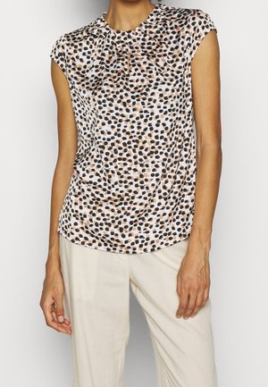 Femme portant un chemisier blanc sans manches avec un motif de points noirs et marrons irréguliers et un pantalon beige clair, debout devant un fond uni.