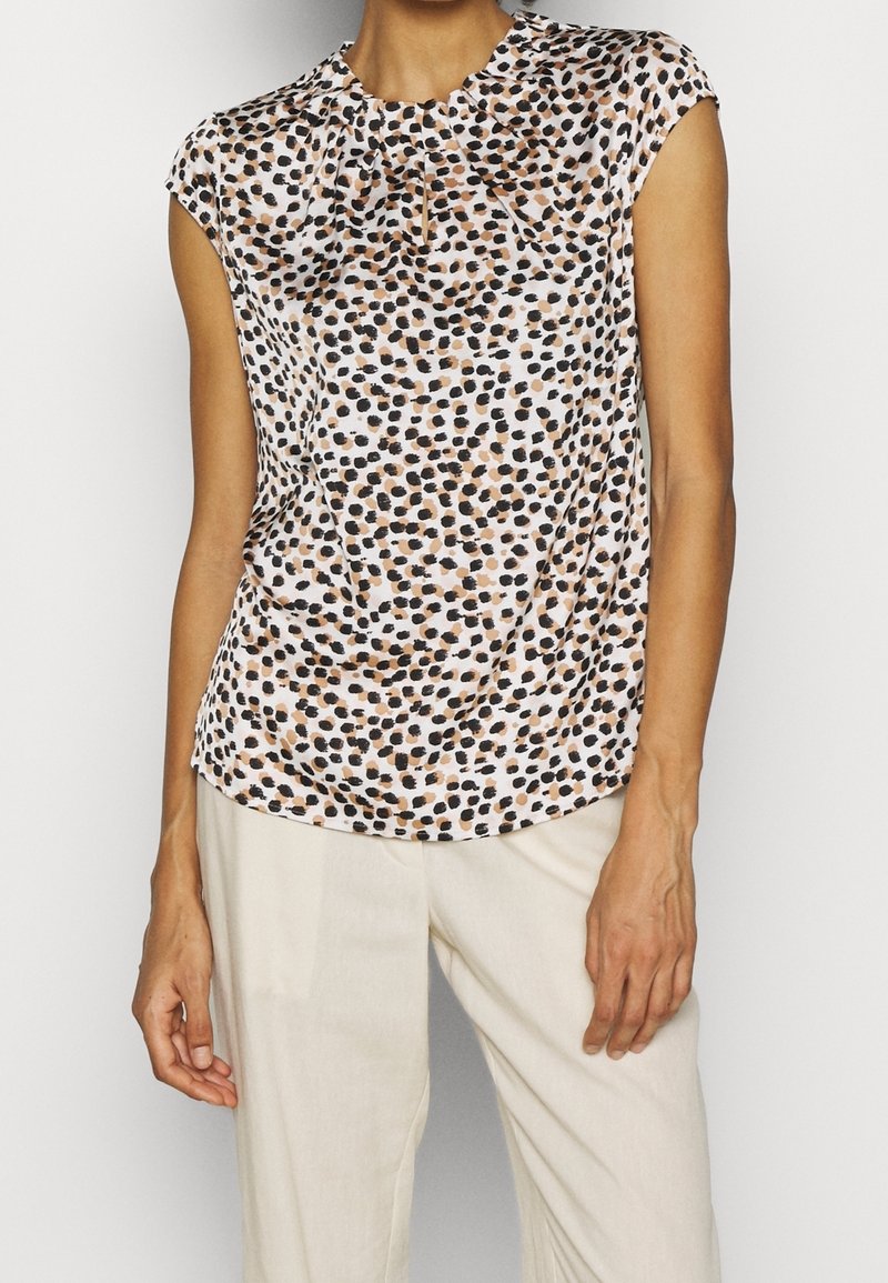 Femme portant un chemisier blanc sans manches avec un motif de points noirs et marrons irréguliers et un pantalon beige clair, debout devant un fond uni.
