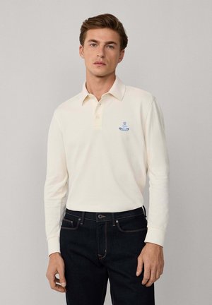 Cremfarbener langärmeliger Polohemd mit Kragen, versehen mit einem blauen gestickten Logo auf der linken Brust, kombiniert mit dunklen Denim-Jeans.