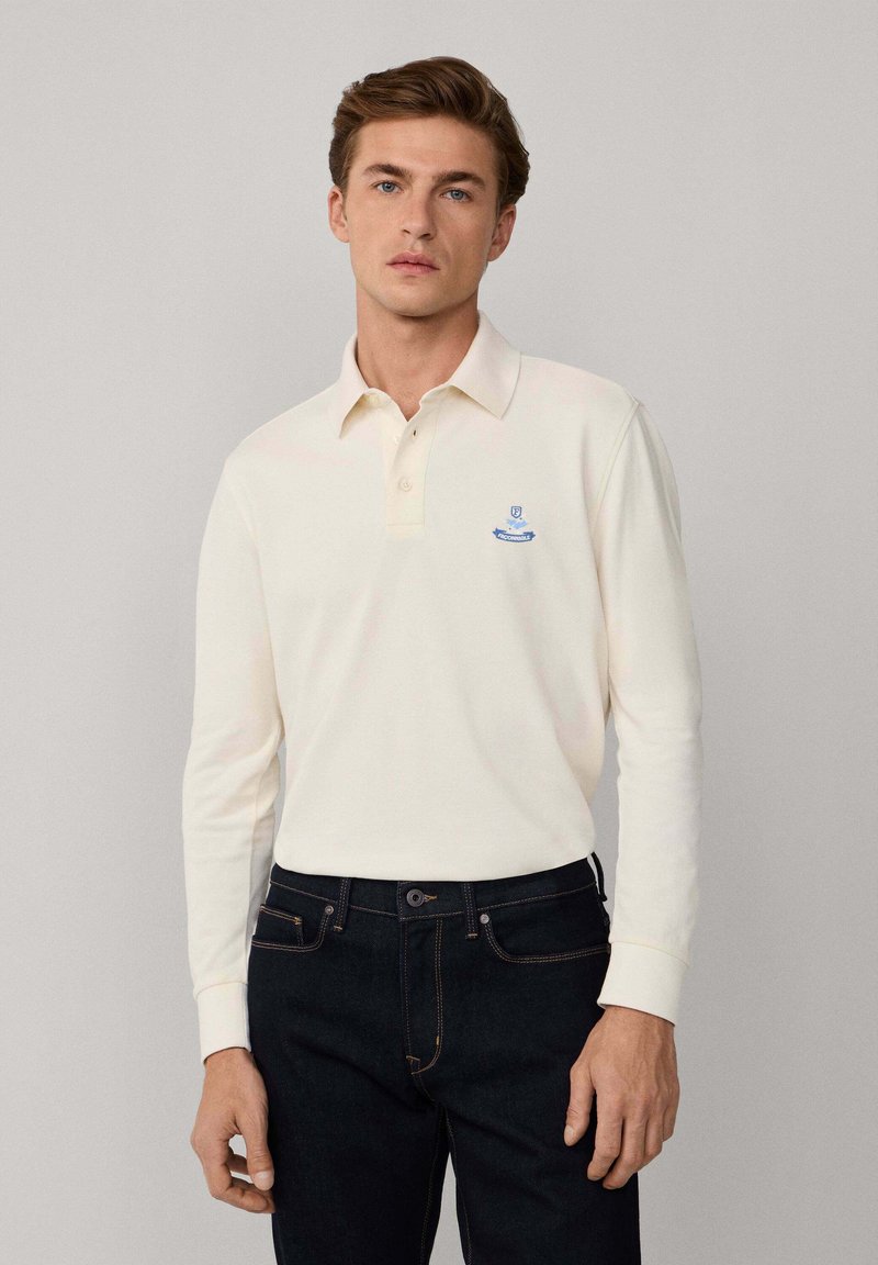 Polo a maniche lunghe crema con colletto, caratterizzato da un logo ricamato blu sulla parte sinistra del petto, abbinato a jeans in denim scuro.