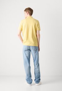Gul bomullspolo med klassisk krage, parret med lyseblå løstsittende jeans og hvite sneakers, sett fra baksiden.