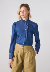 Blå denimbluse med flæsekrave og knaplukning, parret med højtaljede beige bukser med pressefolder og knaplukning.