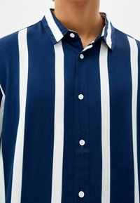 Camicia button-up blu navy con strisce verticali bianche, dotata di colletto e bottoni piccoli. Il materiale sembra liscio e traspirante.