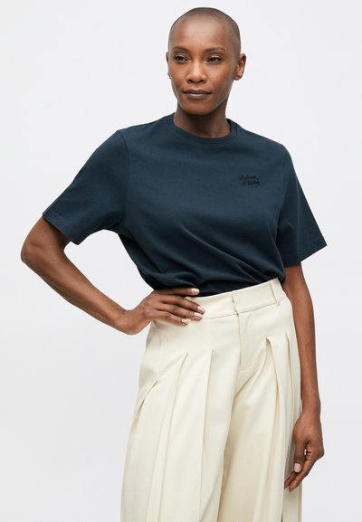 Námořnická modrá bavlněná tričko s krátkým rukávem a kulatým výstřihem, s vyšitým logem na hrudi, v kombinaci s béžovými skládacími kalhotami.