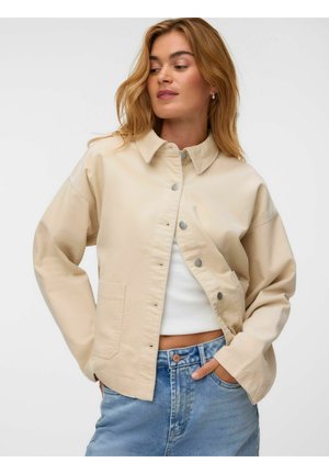 Femme aux cheveux blonds portant une veste beige à boutons sur un crop top blanc et un jean bleu, regardant légèrement sur le côté avec les mains dans les poches.