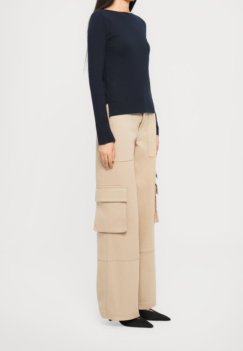 Femme portant un haut noir à manches longues, un pantalon cargo beige à jambes larges avec de grandes poches latérales, et des chaussures noires à bout pointu, debout de côté.