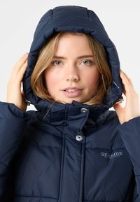 Navy blue puffer jacket with a padded hood, zip closure, snap buttons, and logo detail. Smooth texture with stitched patterns on the fabric.