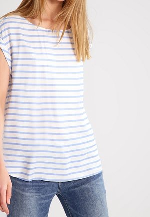 Mujer con una camisa blanca de manga corta con rayas horizontales azul claro y jeans de mezclilla azul, sobre un fondo sencillo.