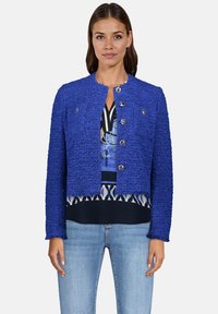 Veste bleue texturée avec un design court, col rond et boutons argentés, portée sur un chemisier à motifs et associée à un jean clair.