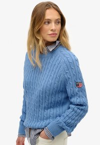 Niebieski sweter z dzianiny w splot warkoczowy, wykonany z teksturowanej tkaniny, z prążkowanym kołnierzem i mankietami, z małą naszywką na górnej części ramienia.