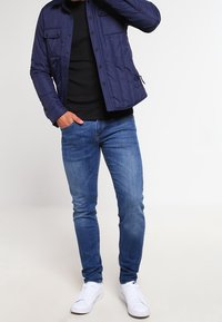 Man i en marinblå vadderad jacka, svart skjorta, blå jeans och vita Nike-sneakers, står med en hand i fickan.